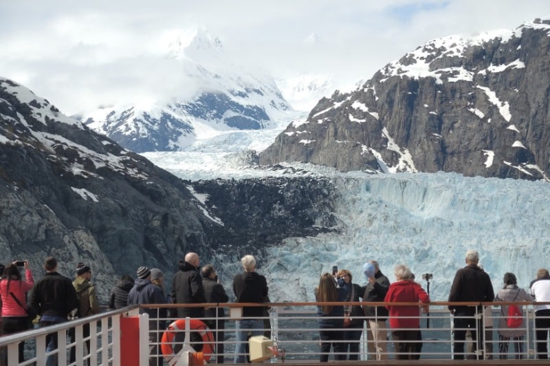 Tourists admire a stunning glacier from a cruise ship deck, surrounded by snowy mountains.